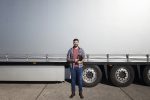 Portrait of middle aged bearded trucker standing in front of truck trailer against grey shiny tarpaulin.